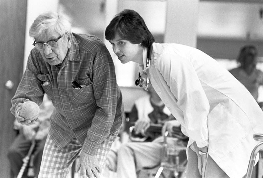 White woman in lab coat stands next to an elderly White man holding a ball.