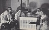 Four seated White men in lab coats look on as a White woman points towards a back lit board.