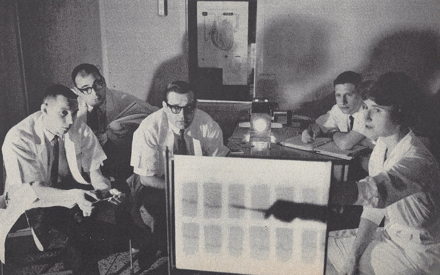 Four seated White men in lab coats look on as a White woman points towards a back lit board.
