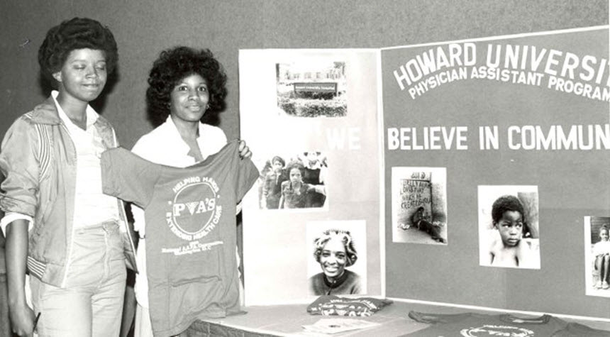 Two African American woman stand next to a poster display, one is holding a shirt.