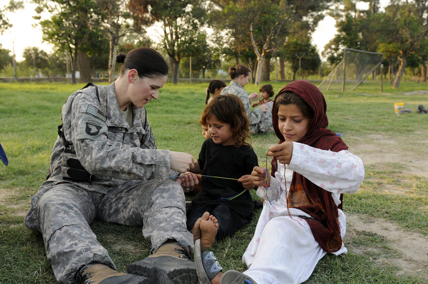 White woman in military camouflage sits in a field with two Middle Eastern girls.