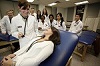 A White woman looks down at woman on examination table, as multiracial group of students look on.