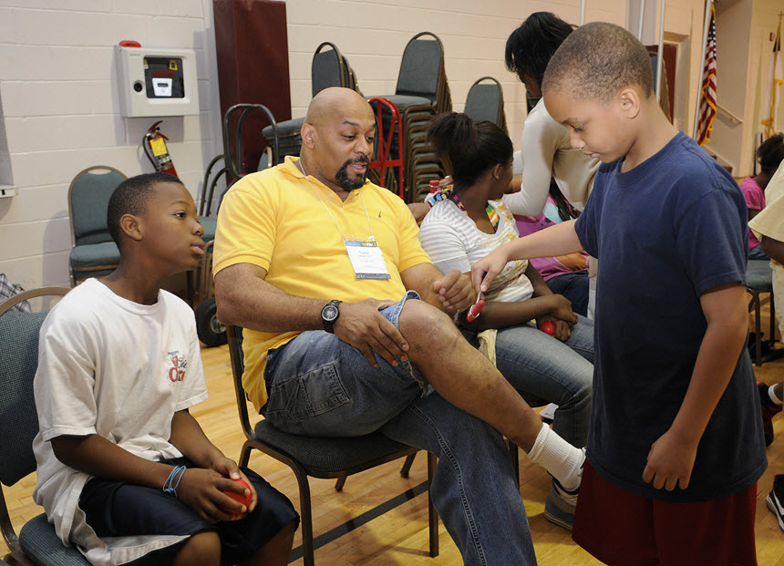 African American boy examines a seated African American man's knee as another boy looks on.