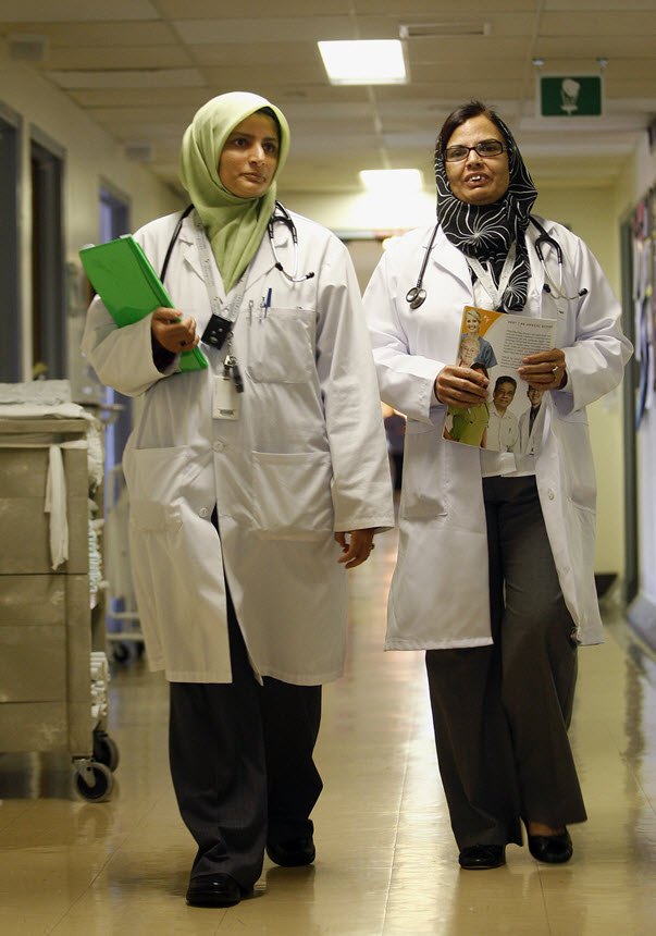 Two South Asian women in lab coats, walk down a hallway towards the viewer.