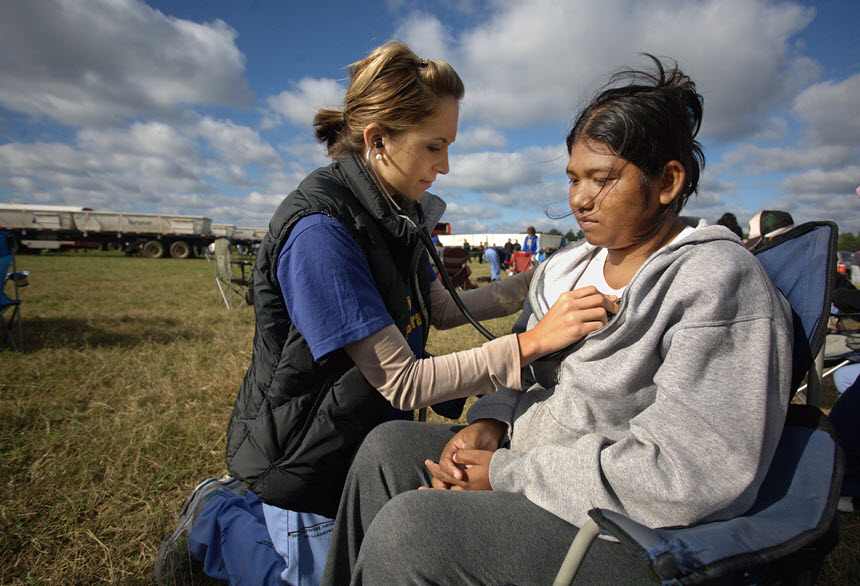 White woman holds a stethoscope to a seated Hispanic woman's chest in a field.