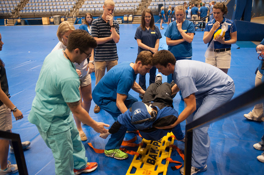 A group of White students lift a mannequin off a stretcher in an auditorium.  