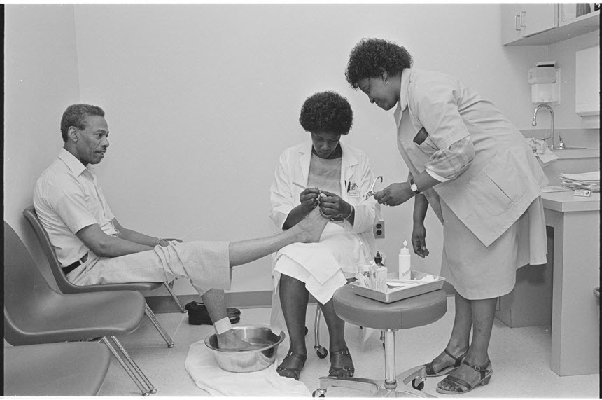 Two African American woman in lab coats tend to the foot of a seated African American man.