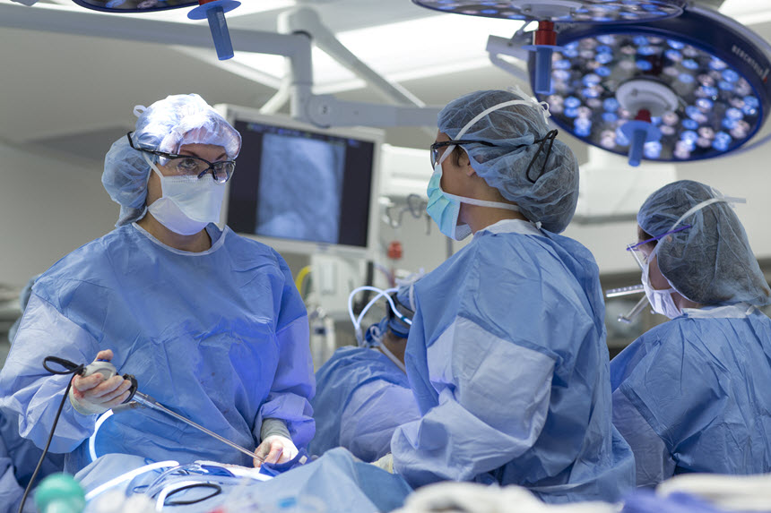 Three people in scrubs perform surgery, one looks off to the right at a screen out of view.