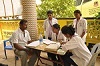 4 South Asian women in lab coats talk to and take notes about a seated man. 