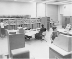 The National Library of Medicine computer room featuring a man seated at a computer terminal in the center of the photograph. In the background, a woman stands in front of a row of tape drives. In the foreground a woman is near some computer equipment. In the top right corner, a person is seated at a desk.