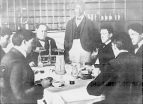 Dr. Wiley, third from left, chief chemist at the USDA, observes five male volunteers at a table with food and drinks. A sixth man, perhaps an assistant and apparently not a volunteer, stands to the left of Wiley.