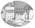 An employee of the FDA sits at a desk and reads the Federal Register. On his desk are over-sized bottles of aspirin and other medications, toothpaste and Pepto-Bismol.
