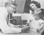 Doctor Adam Metheny, wearing glasses and a white lab coat, leans over a table giving a psychometric tests to a child in the Mental Retardation Clinic of the National Institute of Child Health and Human Development (NICHD). A woman holds the child in her arms.