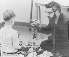 Doctor Milton Shore sits in a chair holding a puppet up in his right hand while a boy looks at doctor Shore.