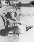 A homeless alcoholic person sits slumped on a park bench. His belongings rest at his side on the bench and on the ground.