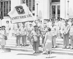 Surgeon General Thomas Parran is reading a speech to Cadet Nurse Corps Director Lucile Petry during the presentation of the Cadet Nurse Corps flag with other Cadet Nurses behind them on steps. A uniformed male holds the flag on the steps directly behind the Surgeon General and Director Petry.