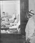 A person wearing protective covering and a plastic mask looks into a room with four prisoner volunteers lying on beds.