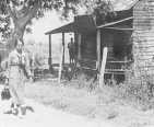  An African American nurse walks past a wooden house; a woman (in silhouette) stands on the porch and watches the nurse.