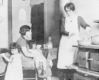 Interior view of a kitchen: a nurse stands at a stove demonstrating proper baby bottle sterilization techniques to a woman seated nearby with a child sitting on her lap; two other small children are present.