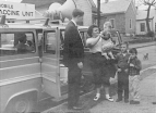 A vehicle with loudspeakers and a sign 'Mobile Vaccine Unit' on its roof stands parked on a street in a residential community. A man stands next to the open door of the vehicle and looks at a woman who holds a baby. Two young boys stand next to the woman.
