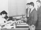 A woman sits at a desk filling out paper work with three men standing in front of the desk.