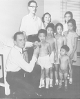 A Public Health Service medical officer at the United States consulate in Hong Kong kneels holding a tongue depressor in the mouth a boy from the Chang Kan Tai family while the other seven family members stand behind them.