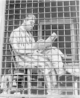 A man in pajamas and robe is sitting in a caged-in porch in the sun, reading a newspaper.