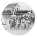 A group of oriental immigrants, many carrying bundles, walk towards the viewer; a building is in the background at the end of a pier
