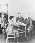 Two men in uniform interrogate a young man. A third man in uniform sits in the back of the room and watches.