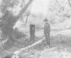 A man wearing a uniform inspects a drip can hanging over a small stream in a wooded area.