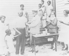 A family stands in front of a building. A physician inoculates the father, while the other family members watch and prepare their own upper arms for the procedure.