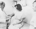 A female survey participant in a hospital gown has her blood pressure taken as part of a complete physical examination by a physician. Another woman stands to the side and watches.