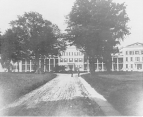 Exterior view of the Marine Hospital, Staten Island, home of the Laboratory of Hygiene for bacteriological investigation.Two large trees are in front of the hospital somewhat blocking the view of the hospital with a road leading between the trees toward the hospital. Two men stand in middle of the road between the trees.