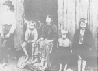 A family, mother and children, all suffering from pellagra. The mother and one of the children sit in the open doorway to their house. The other three children lean against the house.