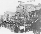 Photograph of a luncheon held outdoors. Rows of men in bowler hats and suits and women in formal wear and hats are shown at long tables positioned on a street in San Francisco. Several buildings appear in the background, including one, with a sign [G]eorge J. Ellington on its roof.