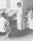 Three researchers in a laboratory. Two are seated on stools while the third one stands. All are wearing white lab coats.