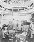 An operating room during a surgery. Several people dressed in hospital scrubs surround the operating table. A nurse stands in the side of the table with their hand behind their back. Another person is seated on a stool in front of an instrument panel. A person looks down on the the operating room from an observation room.