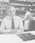 Dr. Robert C. Gallo seated at a table in a laboratory with his arms crossed. Behind Dr. Gallo, a research works in the laboratory.