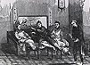 Four Chinese men in traditional dress smoke opium on a bench