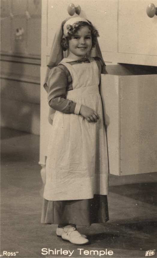 White female child-actress (Shirley Temple) dressed as a nurse and looking at the viewer.