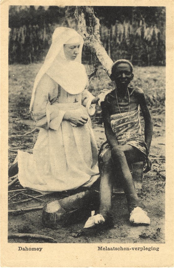 A White female religious nurse kneeling and tending to a thin African young woman.