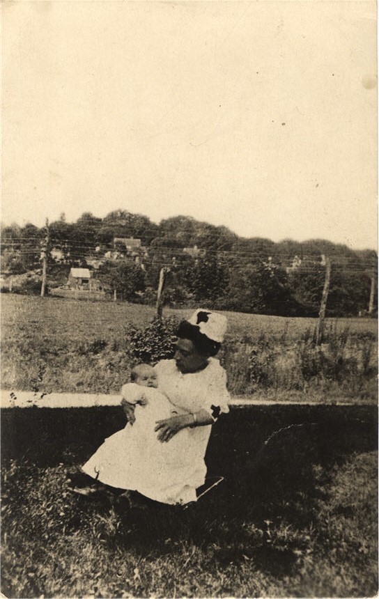 An African American female little person nurse holding a White infant and sitting outside.