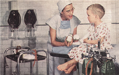 A White female nurse anesthetist talking to a White boy sitting on an operating table.