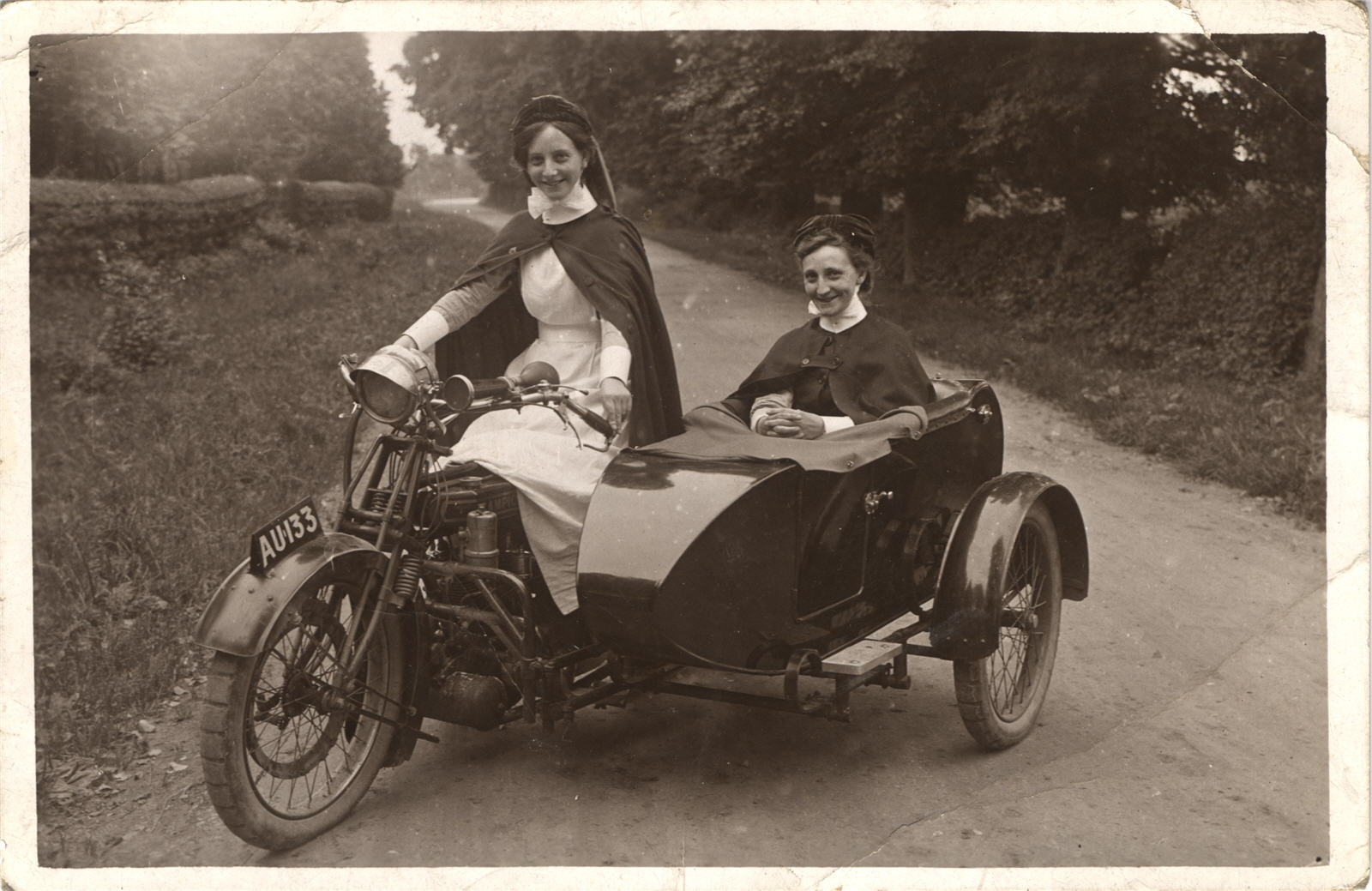 Two White female nurses in a motorcycle with sidecar. Both wear white uniforms under their cloaks.
