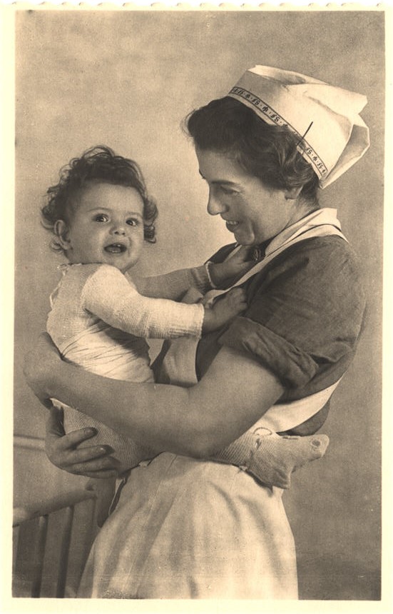 A White female nurse holding a White baby, who is smiling at the viewer.