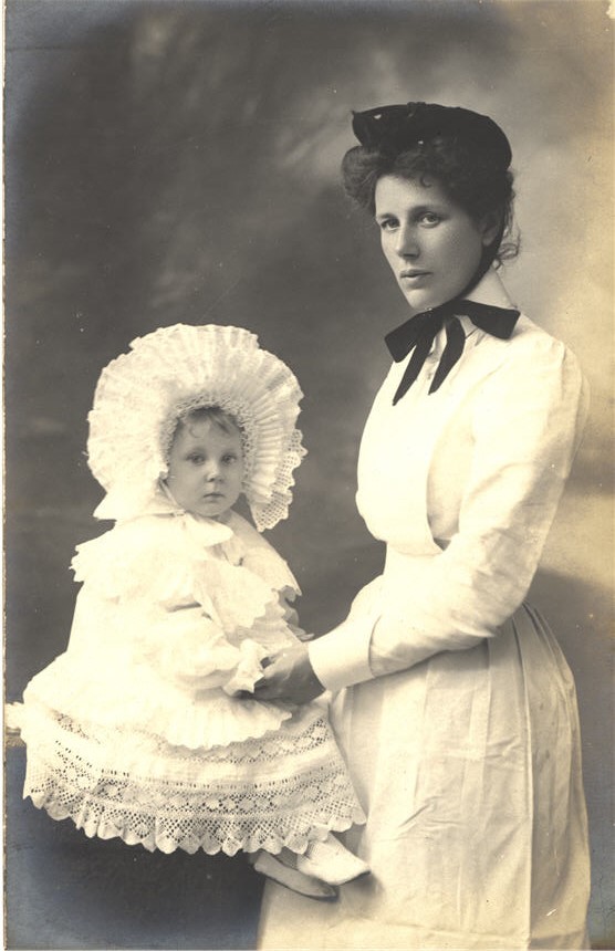 A White female nursemaid in white uniform, standing with a White girl white gown.