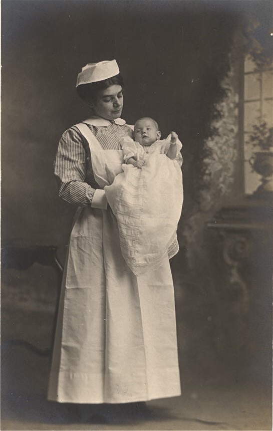 A White female nurse holding a White infant. The nurse looks down at the baby.