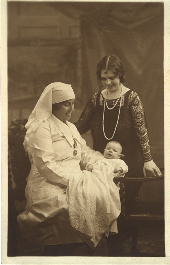 A White female nurse sitting and holding a White baby. Another White woman stands behind.