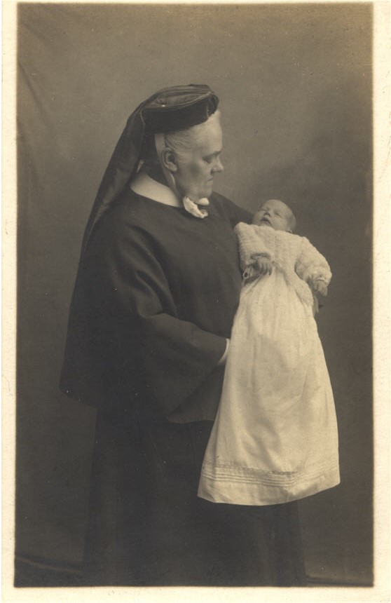 An elderly White female nurse in black, holding a White infant. The nurse looks at the baby.