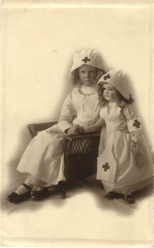 A White girl dressed in a white nursing uniform sits next to a standing nursing doll.